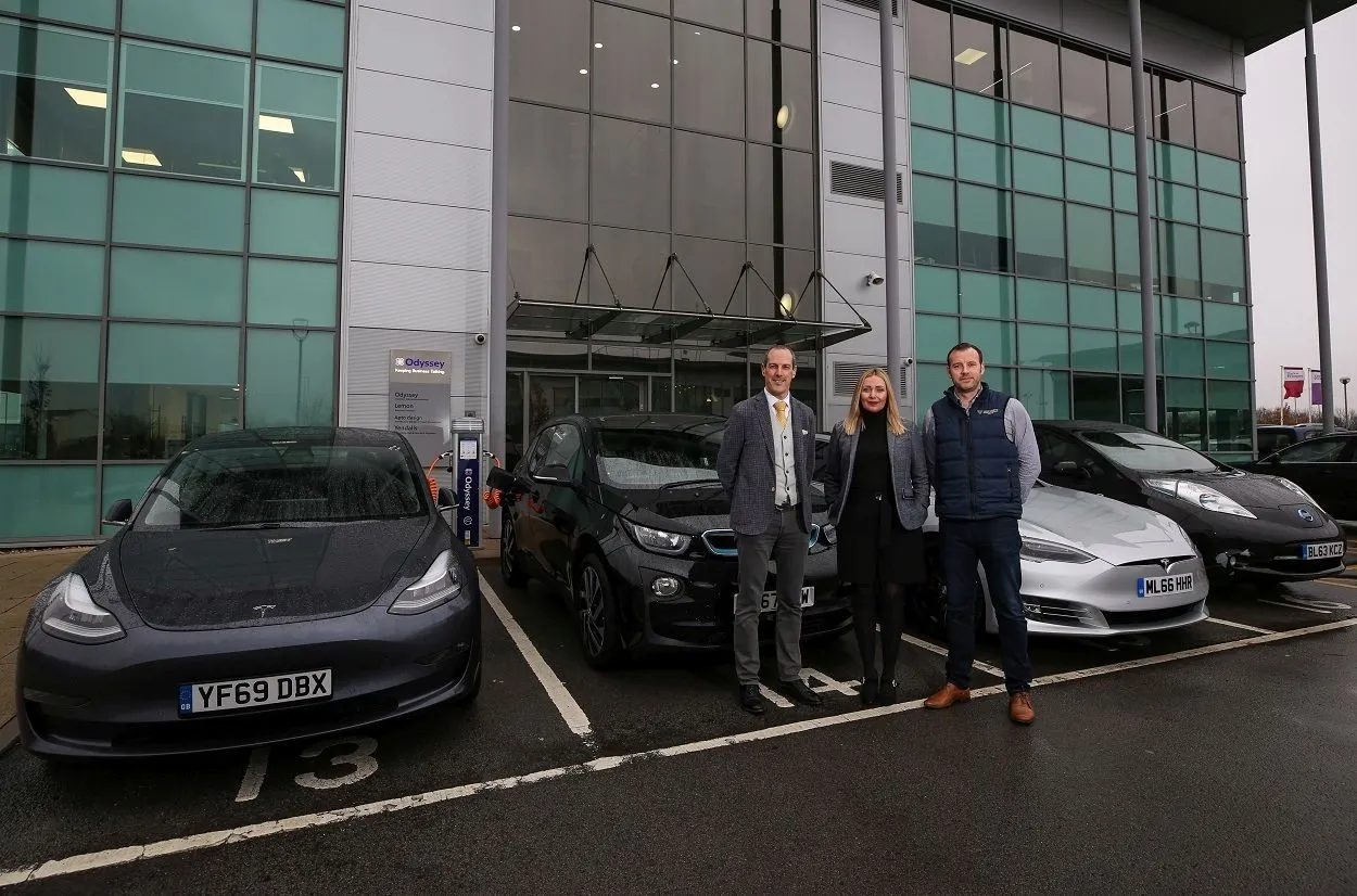 Three people from Odyssey standing beside electric cars parked at the charging point outside the Preston Farm offices