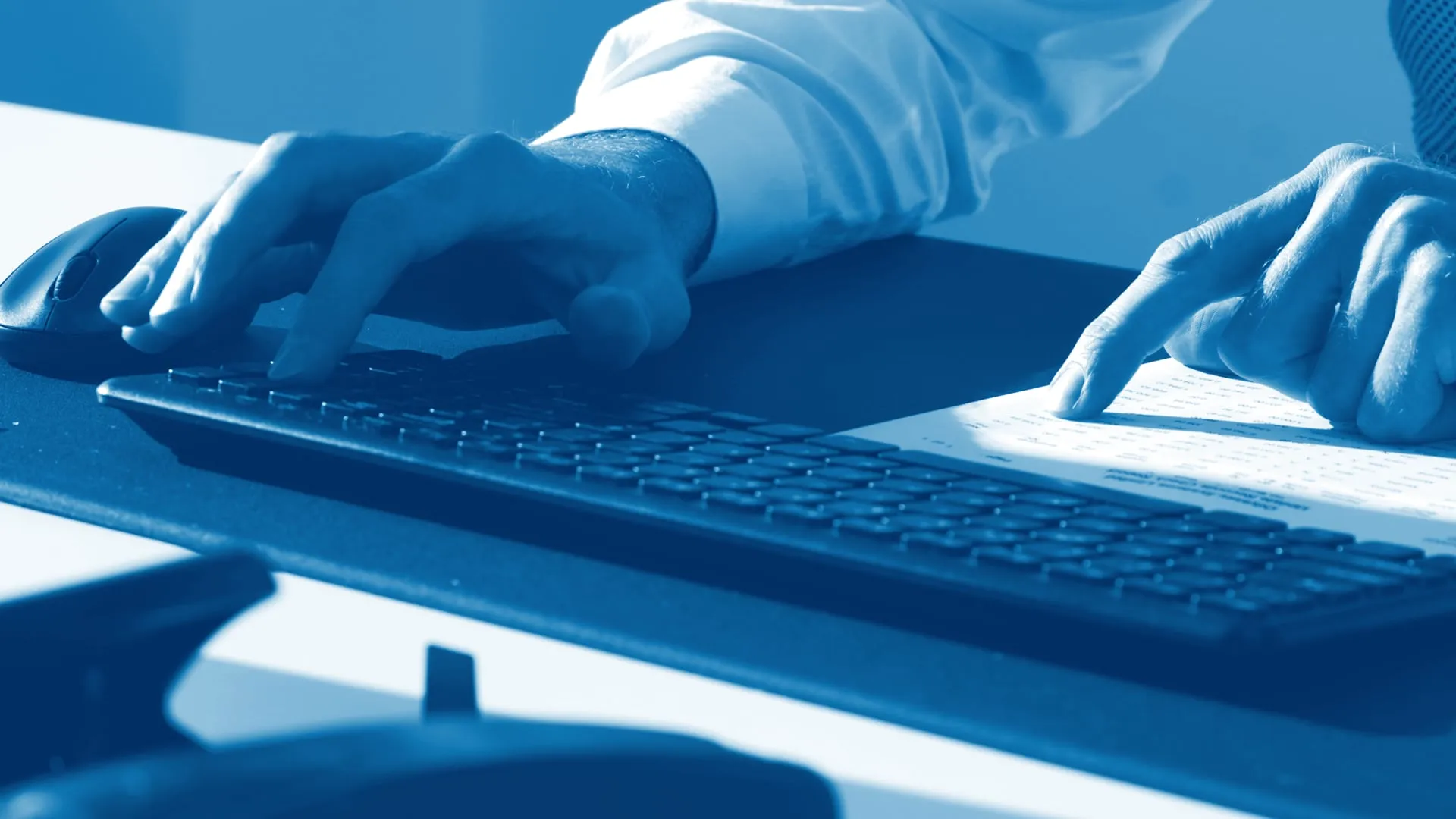 Hands at a keyboard and mouse, one hand following a line on a printed document