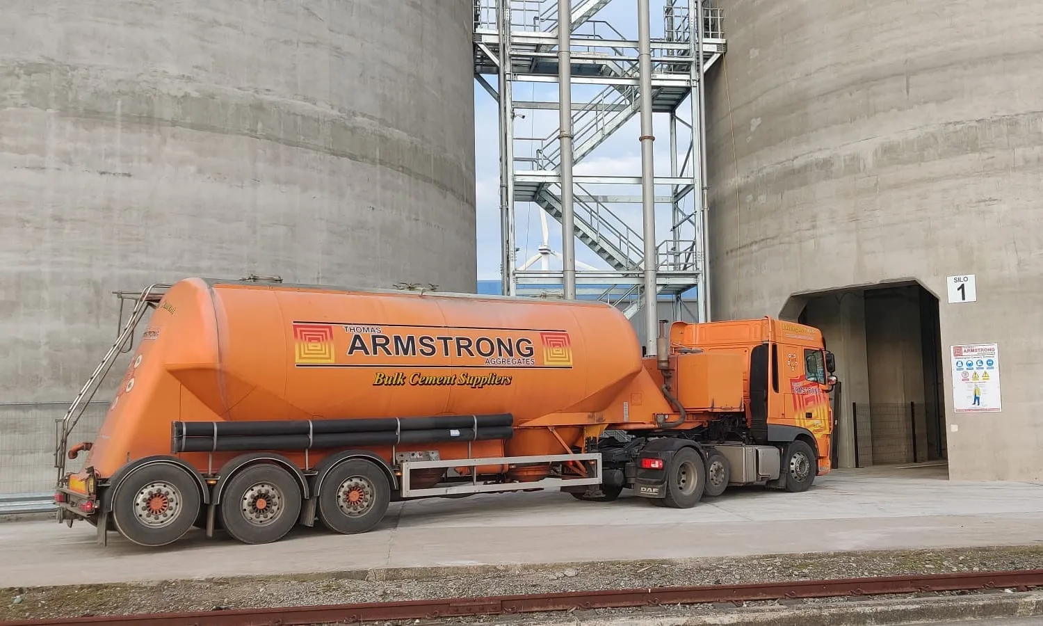 An orange Thomas Armstrong Aggregates bulk cement tanker parked by Silo 1 between two concrete silos